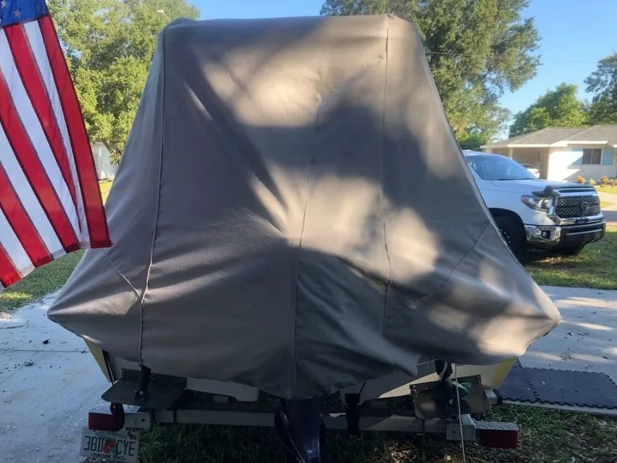Boat on a trailer covered in a tan tarp; American flag to the left, truck and house in the background.