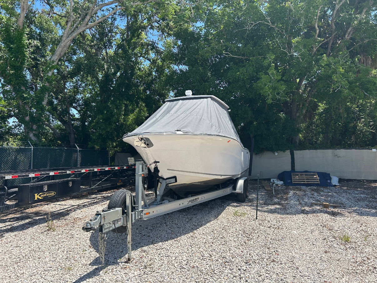 Boat on trailer, covered in gray tarp, parked on gravel. Trees and fence in the background.