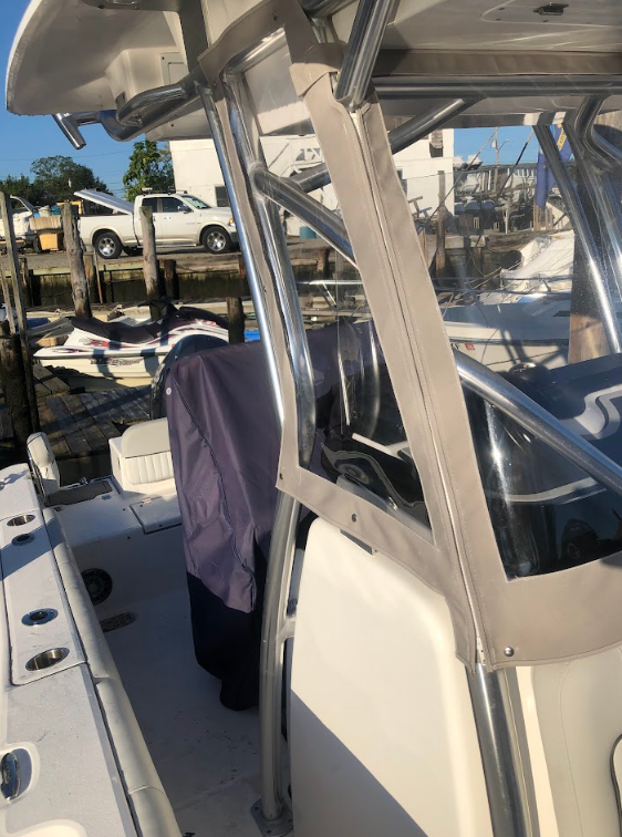 Boat's helm with canvas, stainless steel frame. Beige and white deck. Harbor in background with other boats and a truck.