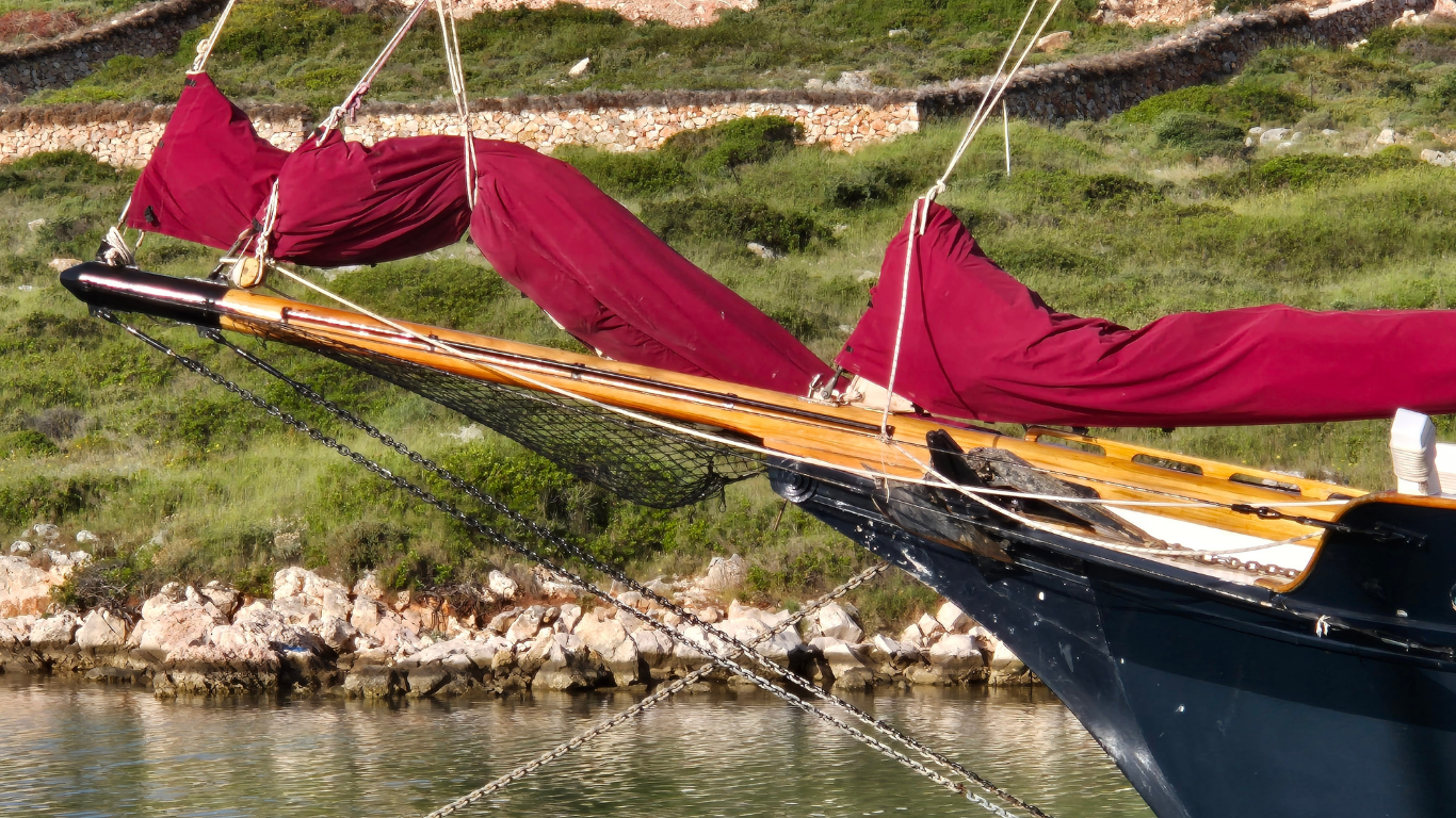Bow of a dark blue sailboat with red sails, near green grass and stone wall.