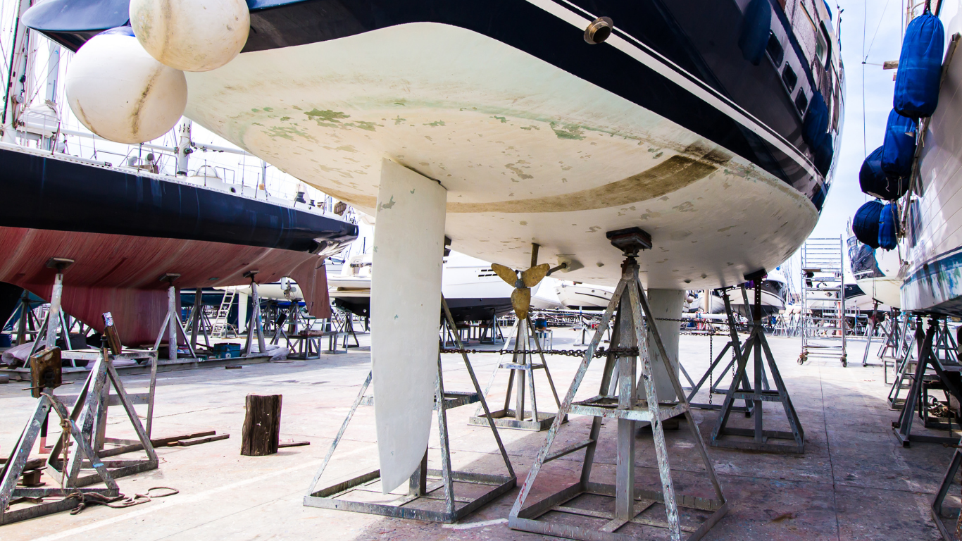 Boat hull in a boatyard, propped up on jacks for maintenance; other boats in the background.
