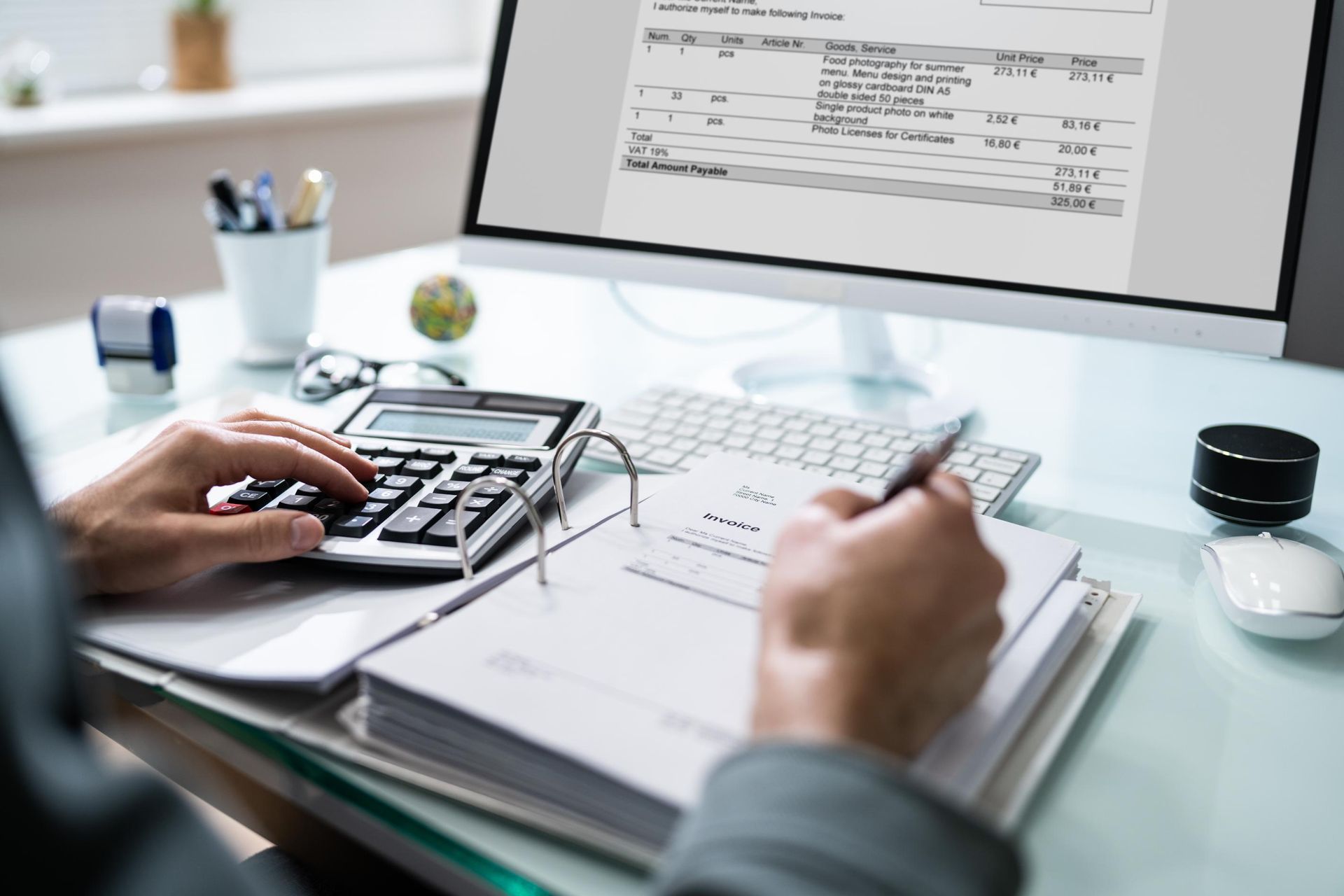 Person calculating financial data with a calculator, computer, and paperwork at a desk.