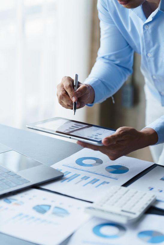 Man in blue shirt pointing at tablet displaying charts, surrounded by financial documents and laptop.