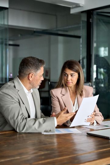 A man and a woman in suits review documents at a table in an office.