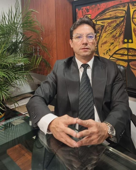 Un hombre con traje y corbata está sentado en una mesa con las manos juntas.