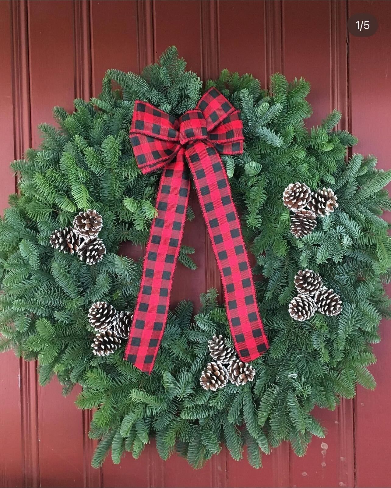 A christmas wreath with pine cones and a red and black checkered bow is hanging on a red door.