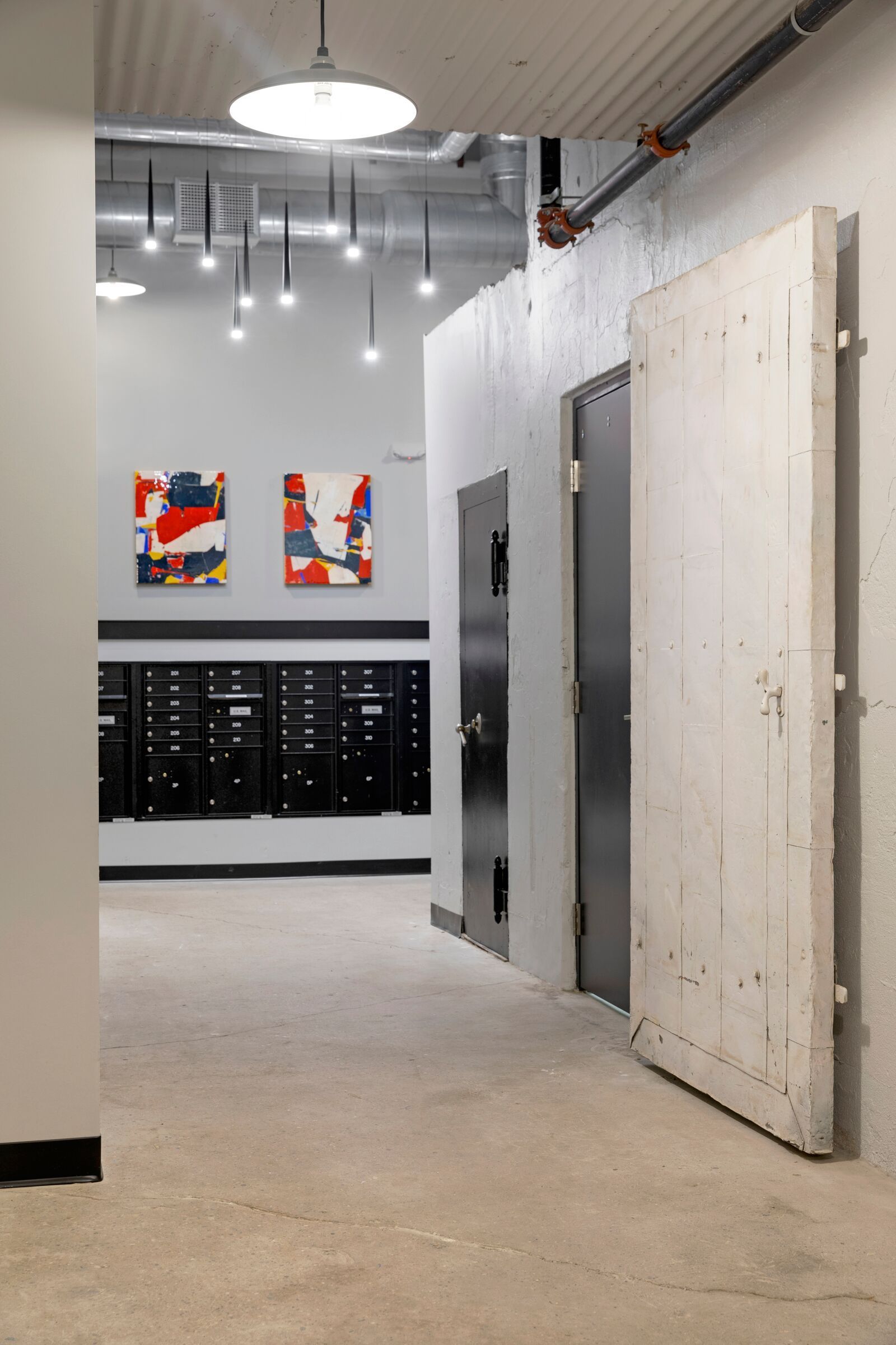 Interior hallway in a multifamily building featuring mailboxes, abstract art, exposed pipes, and a large white wooden door at The Jax in St. Paul, MN.