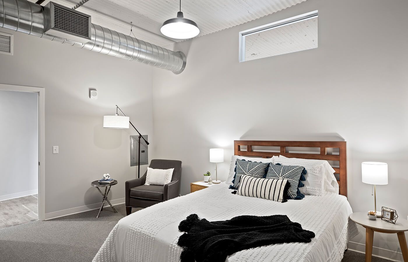 Modern apartment bedroom with white bedding, wooden headboard, and exposed industrial ceiling ducts at The Jax in St. Paul, MN.