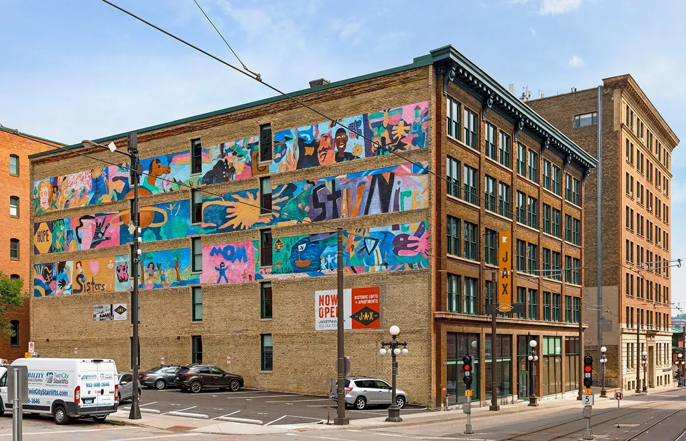 Brick building with vibrant mural and cars parked nearby. Street and sky visible.
