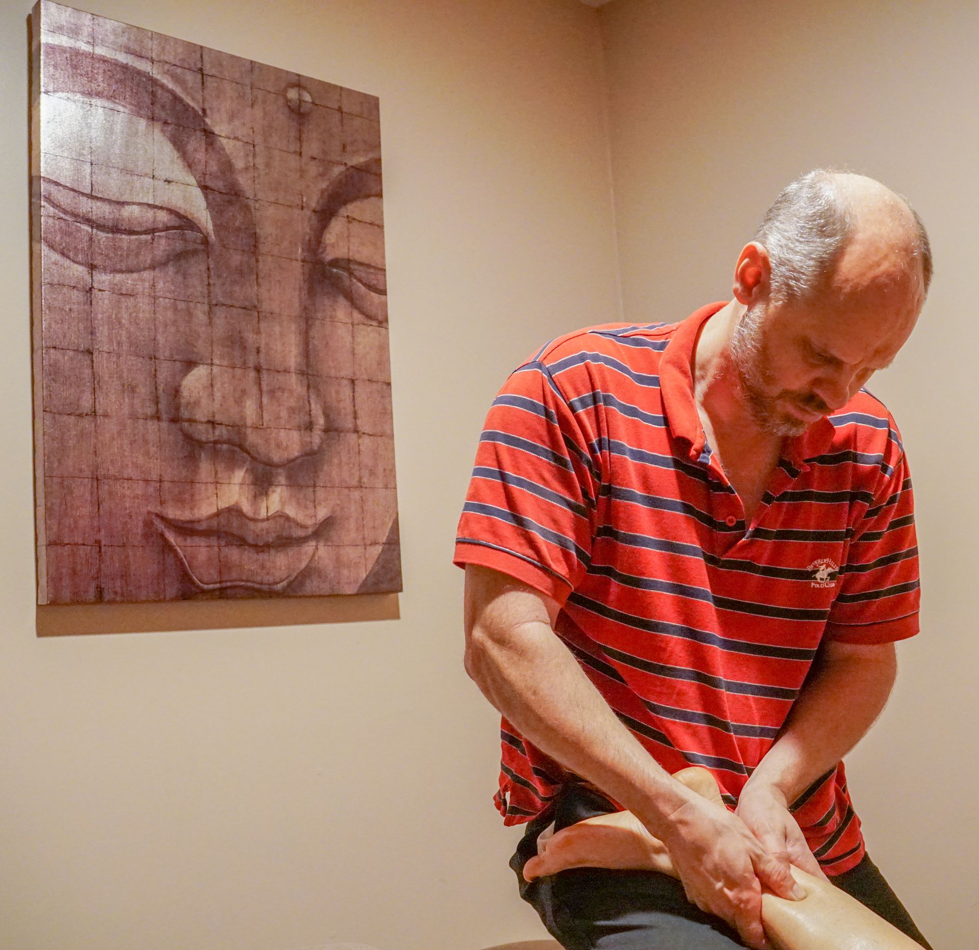 Man in a red striped shirt seated beside a wall portrait, looking down and holding his leg.