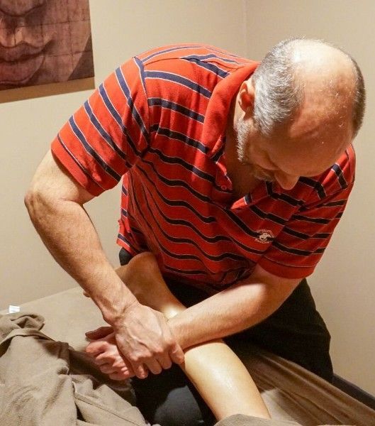 Man massaging another person's calf on a treatment table in a room, wearing a red striped shirt