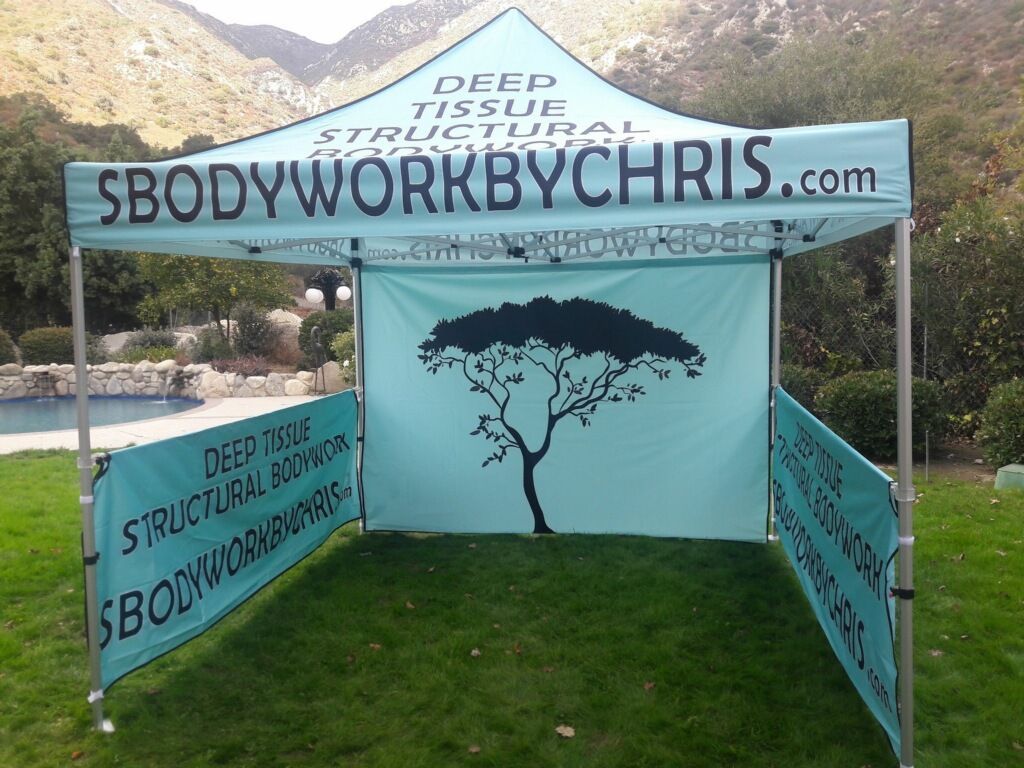 Booth tent with turquoise banners reading “SBODYWORKBYCHRIS.com” at an outdoor market with mountains behind.