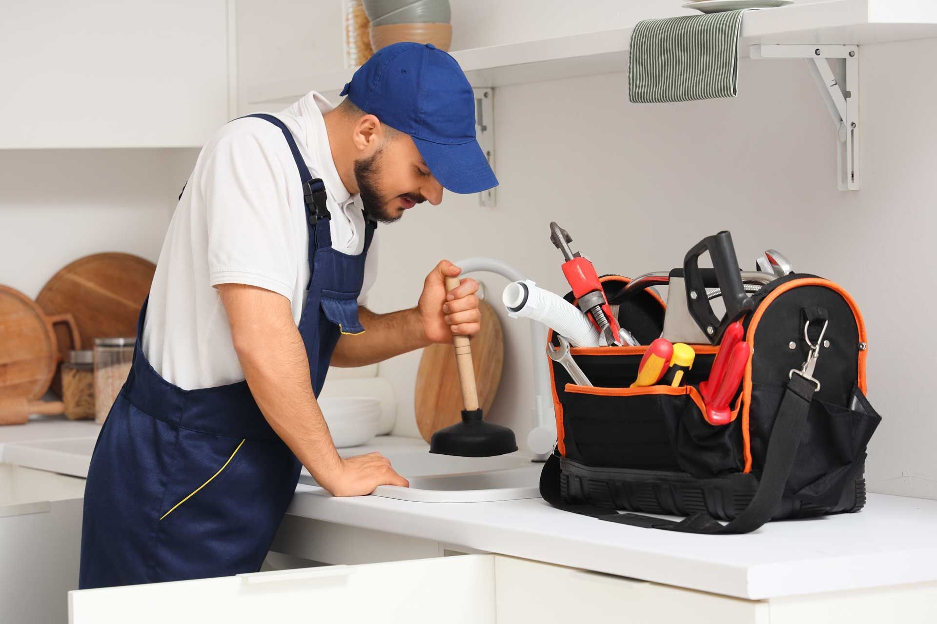 Plumber in blue overalls and cap with a plunger, examining plans near his toolbox in a kitchen.