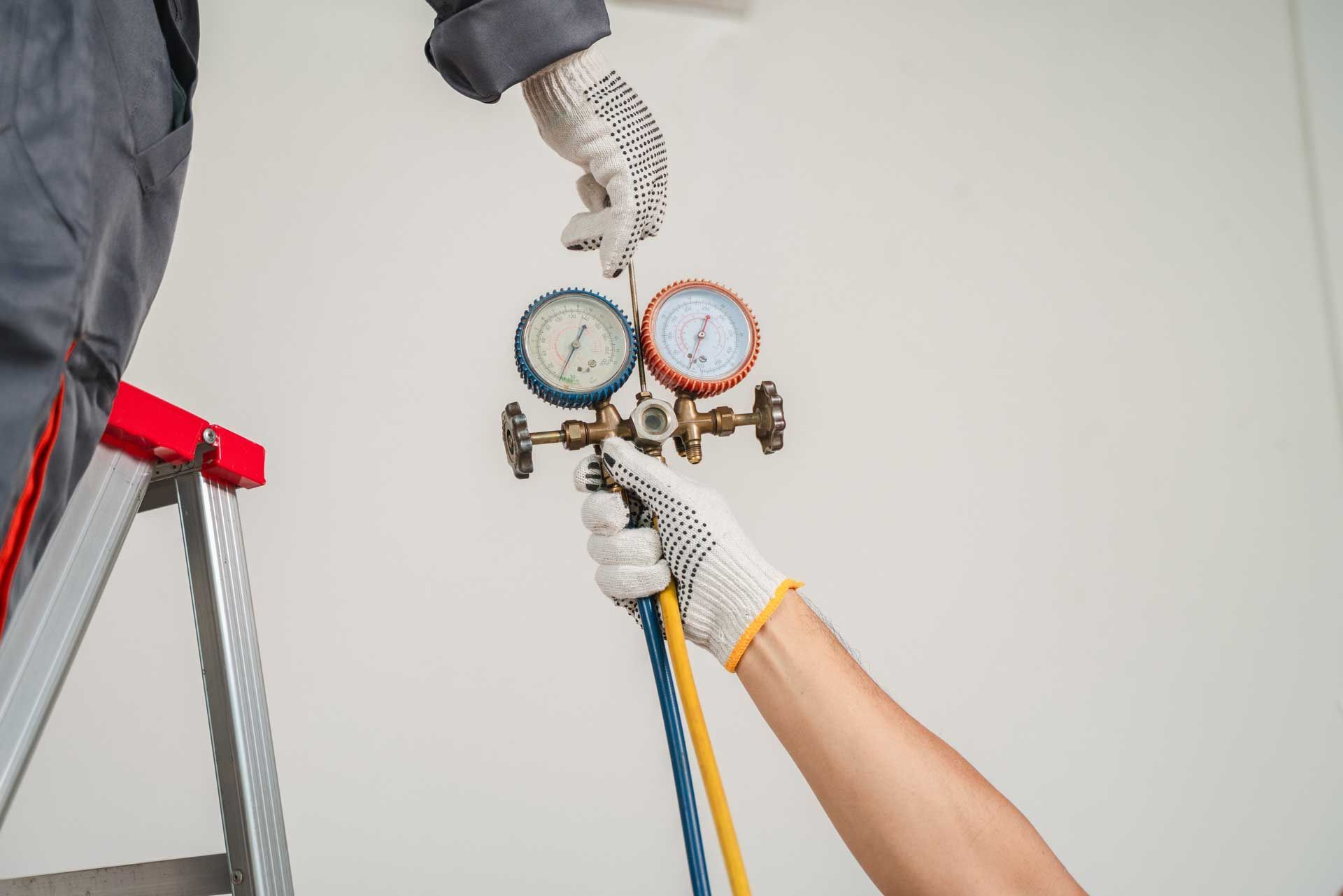 Two workers connecting gauges to an HVAC unit, on a ladder, against a white wall.