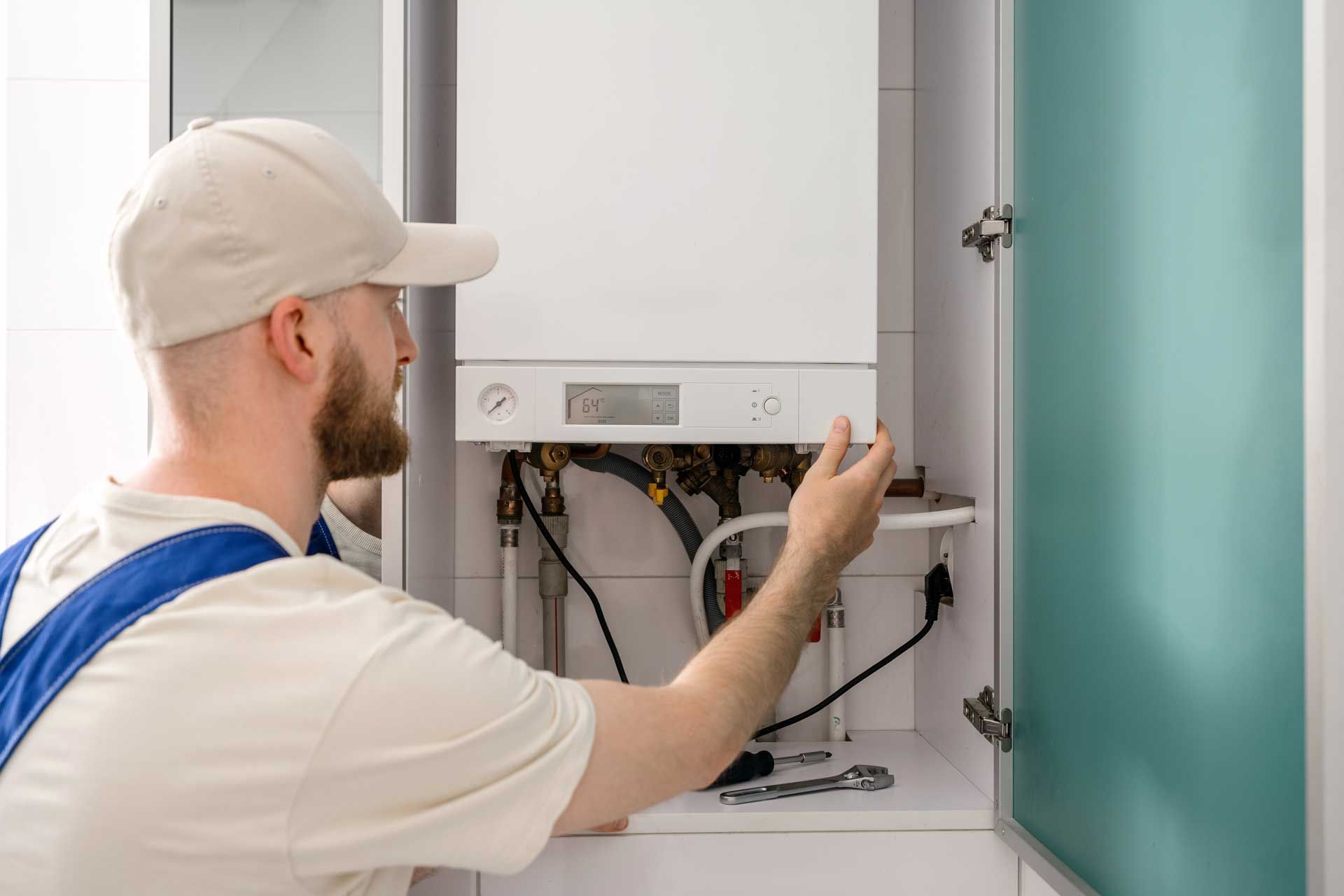 A person in work clothes inspects a white heating unit in a cabinet.