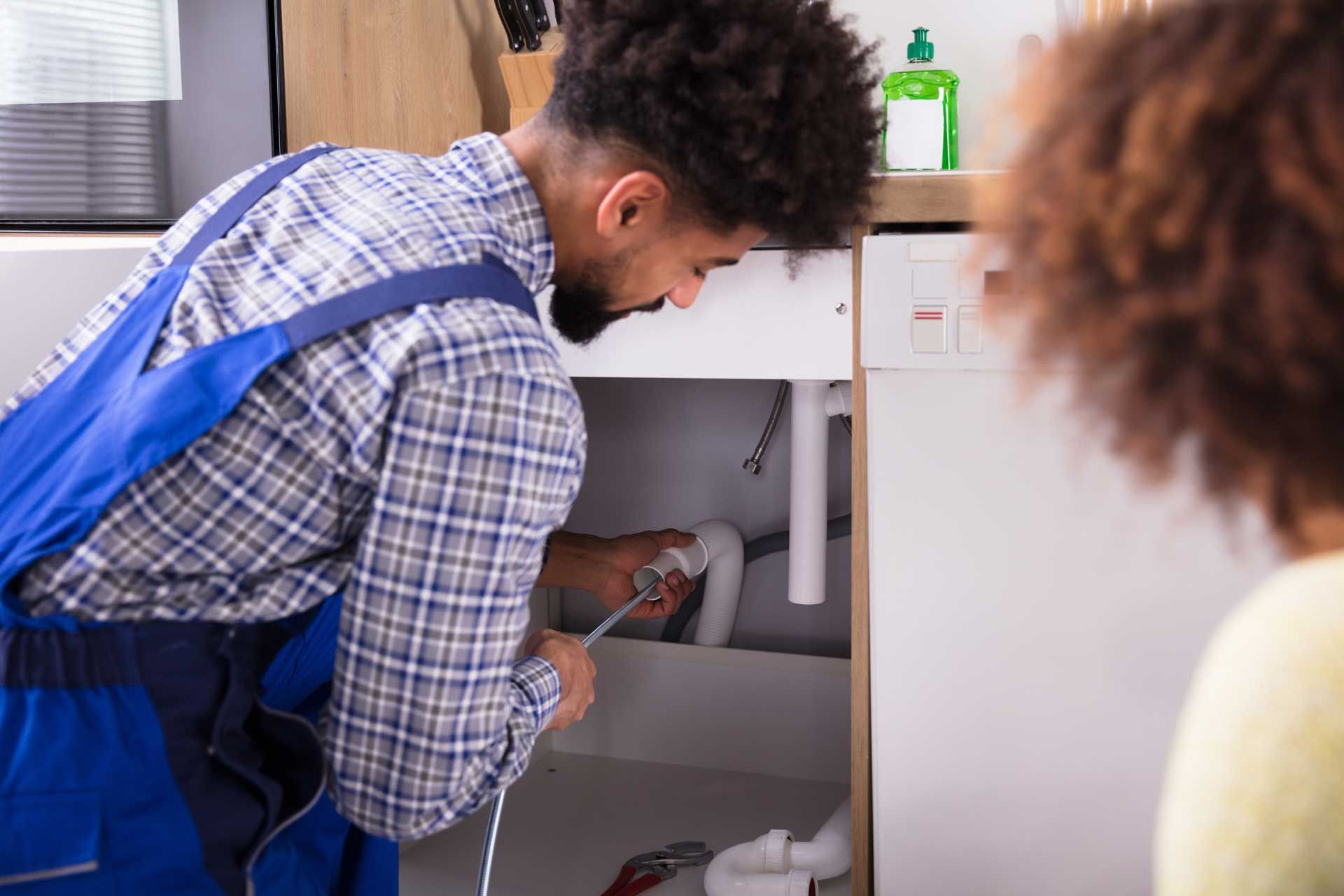 Plumber in blue overalls and plaid shirt works under a sink, another person watches.