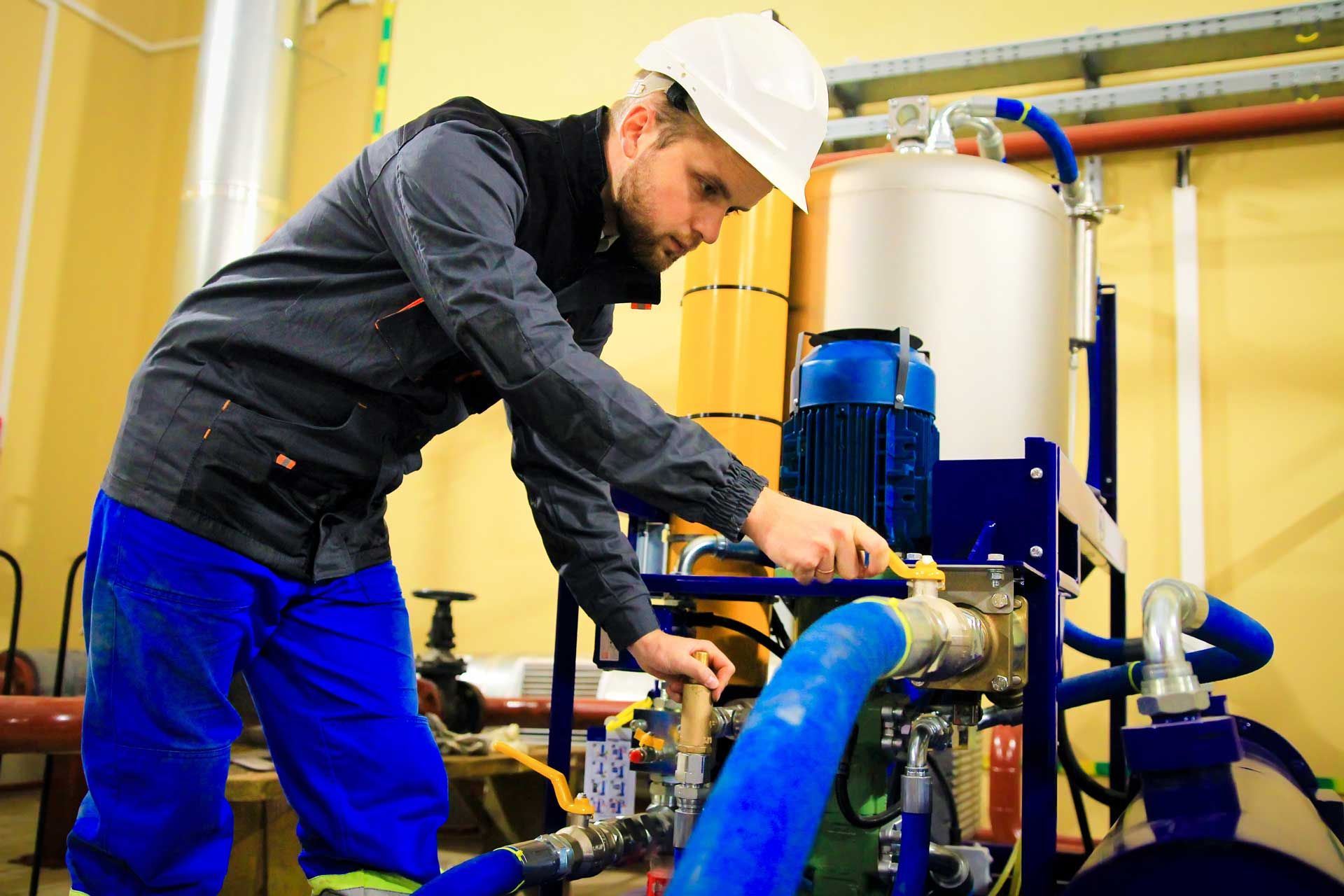 Man in blue work clothes and white hard hat adjusting machinery in a utility room.