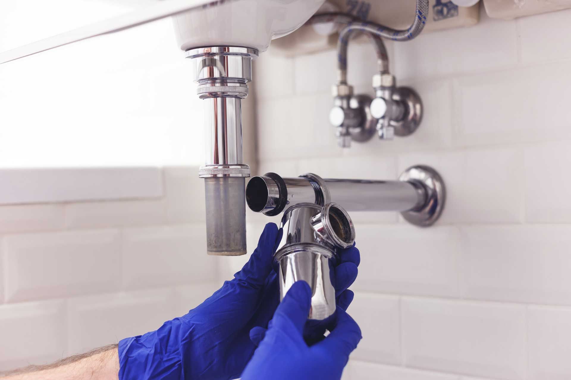 Hands in blue gloves assembling a chrome P-trap under a sink. White tile backdrop.