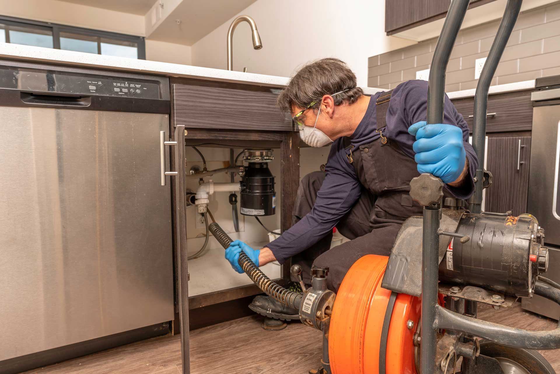 Plumber using a drain snake under a kitchen sink, wearing gloves and mask.