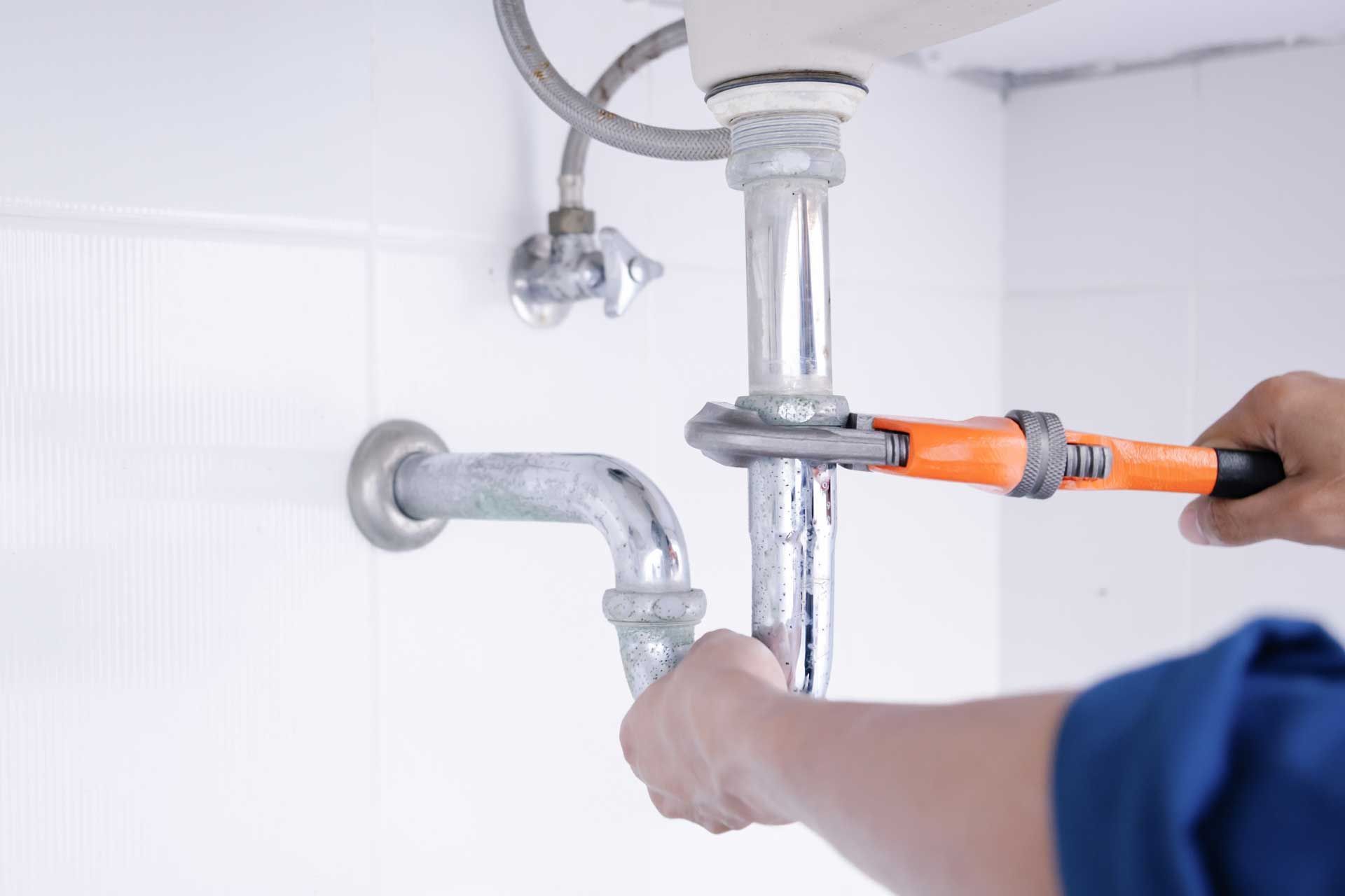 Plumber using an adjustable wrench to work on a chrome pipe under a sink.