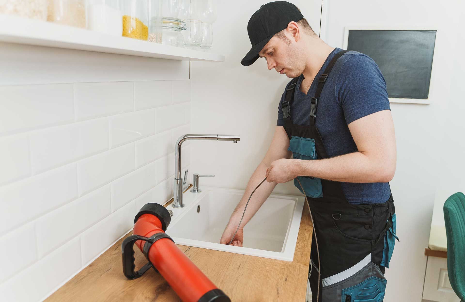 Plumber in a kitchen, using a snake tool to unclog a white sink. He wears a hat and overalls.