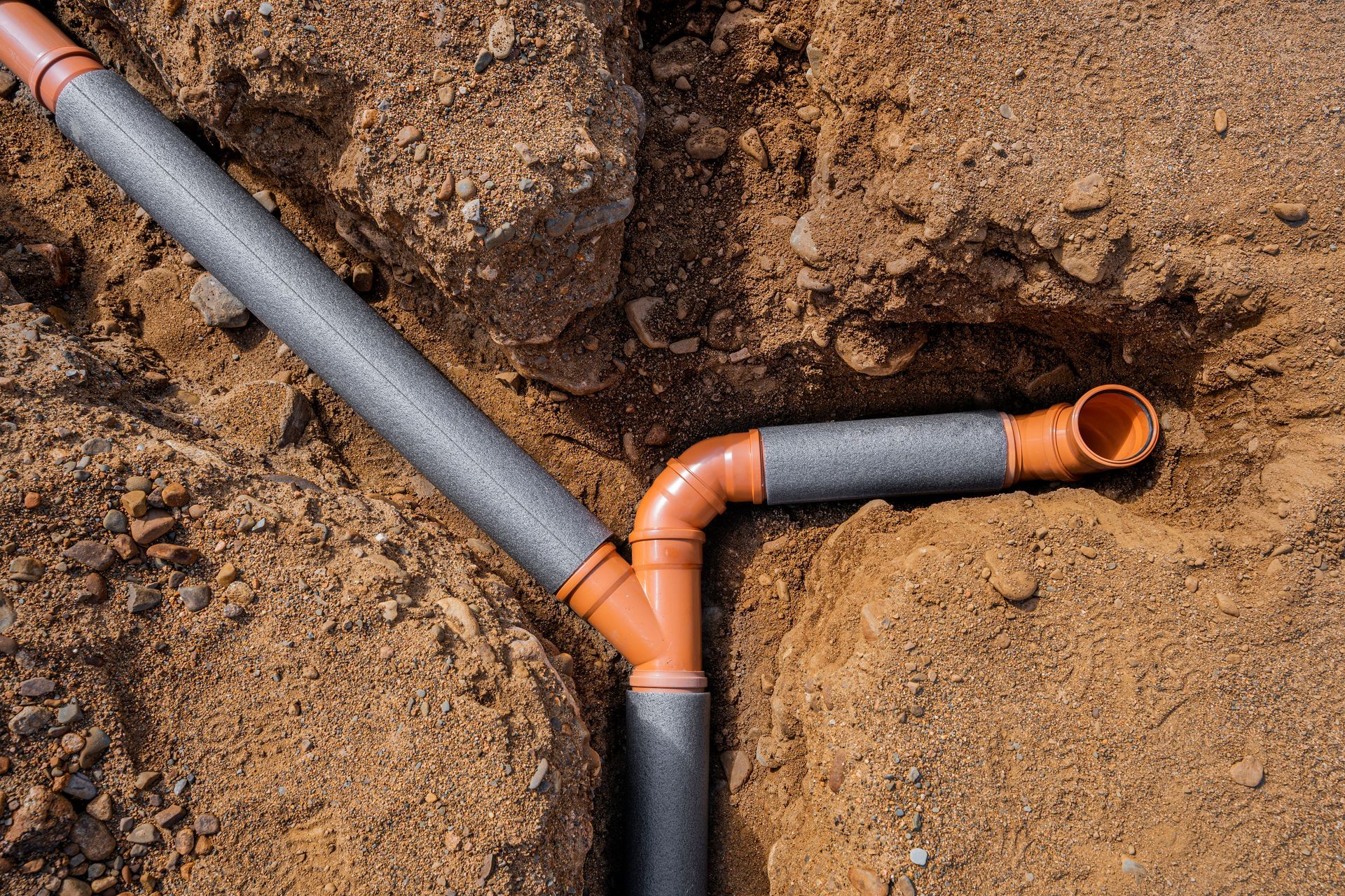 Orange and gray drainage pipes installed in a trench dug into brown soil.