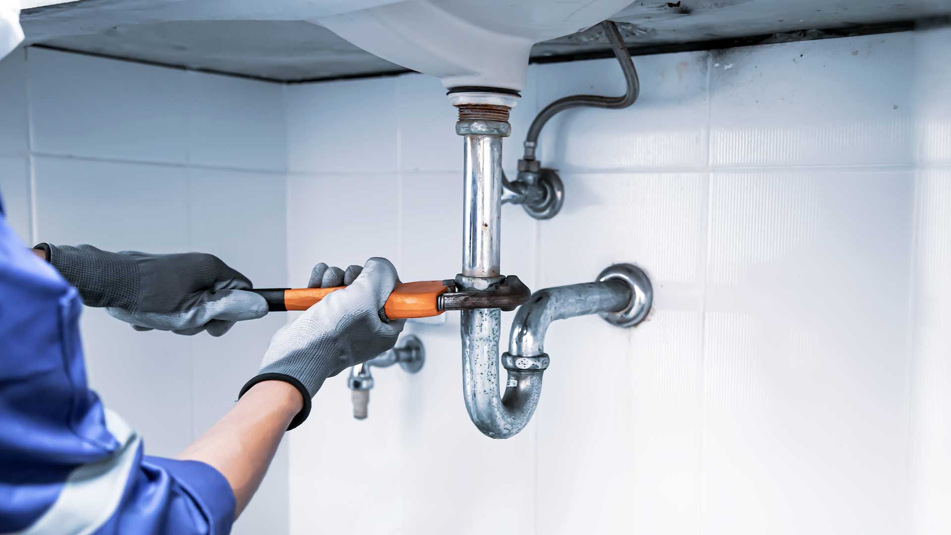 Plumber using a wrench on a bathroom sink's drain pipe, hands in gloves, white tile background.