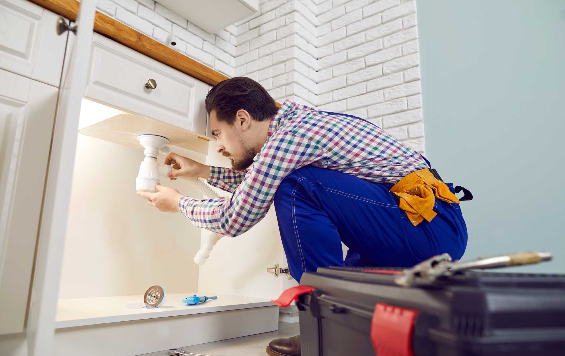 Plumber working under a kitchen sink, holding a pipe. Wearing blue overalls and plaid shirt.