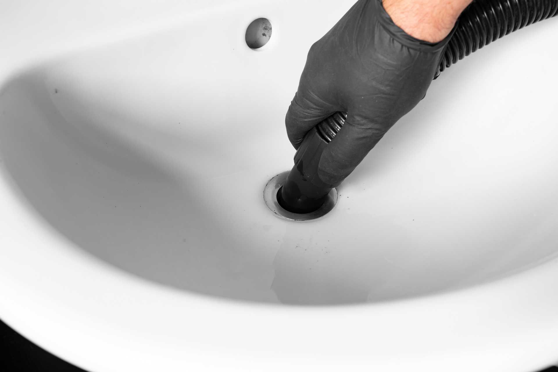 Gloved hand holding a black hose into a white bathroom sink drain.