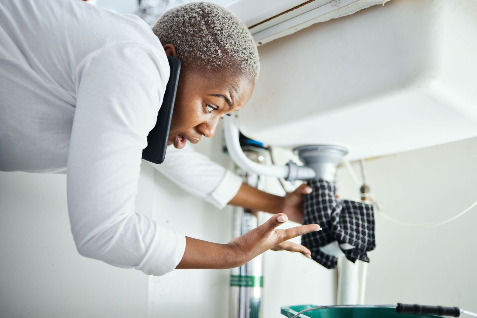 Person on the phone, looking concerned while working on plumbing under a sink.
