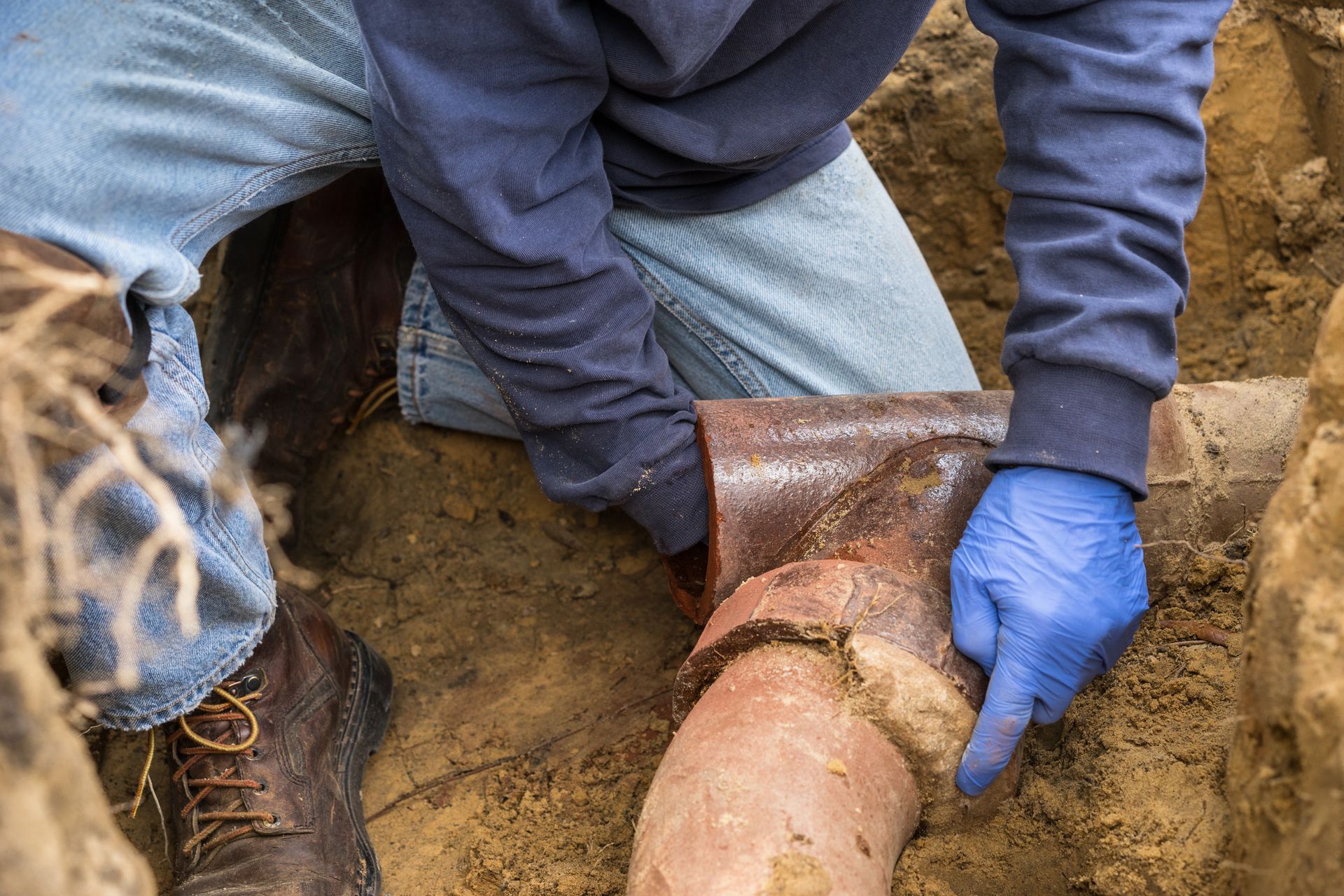Person wearing blue gloves pointing at a broken underground pipe in a dirt trench.