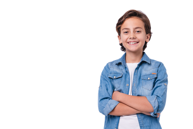 A young boy in a denim shirt is standing with his arms crossed and smiling.