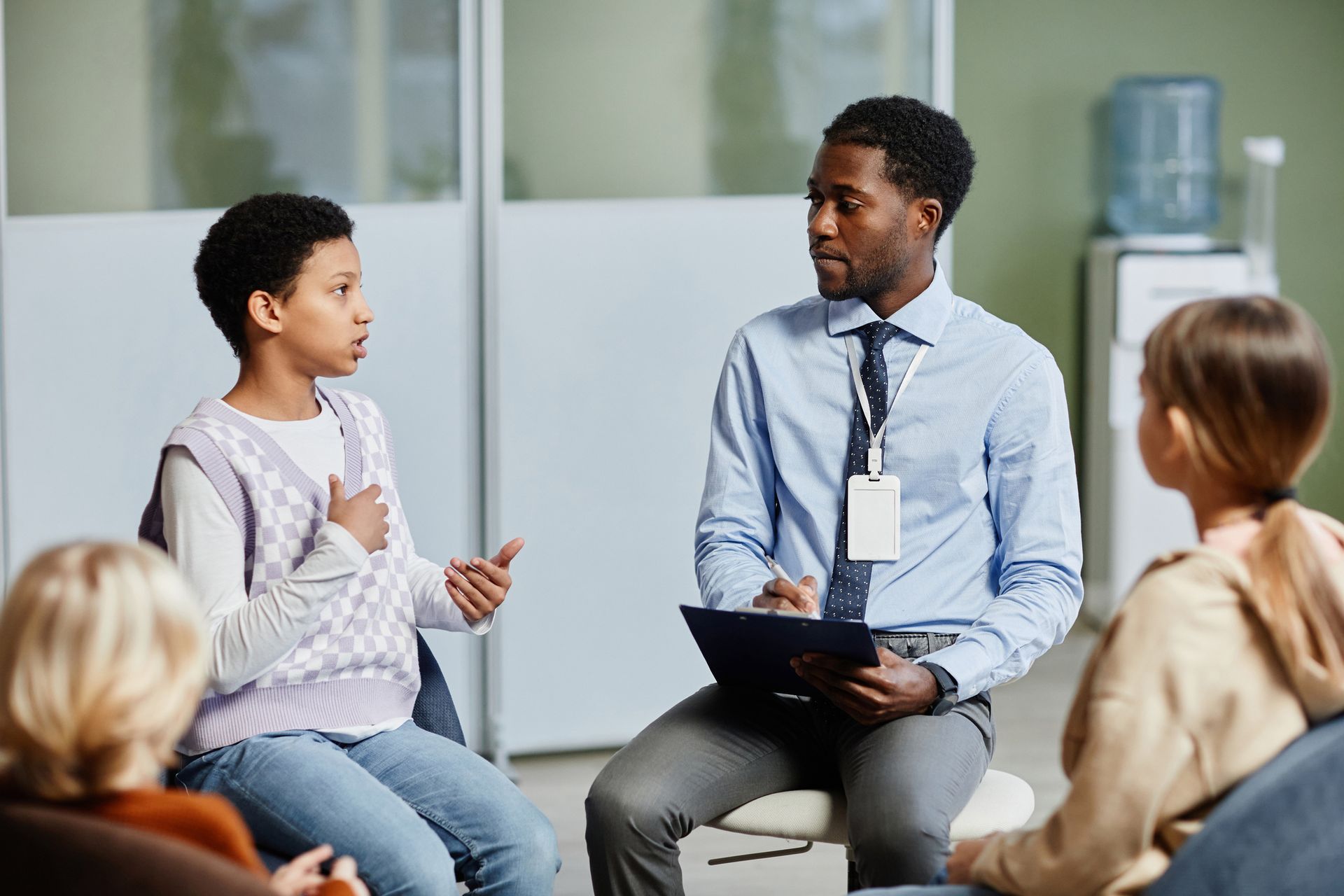 A group of people are sitting in a circle talking to a man holding a clipboard.