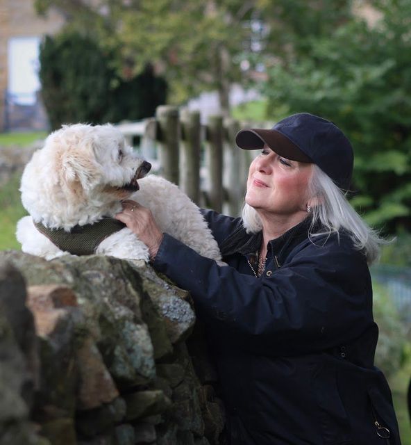 Woman in a cap smiling, petting a fluffy white dog on a stone wall outdoors.