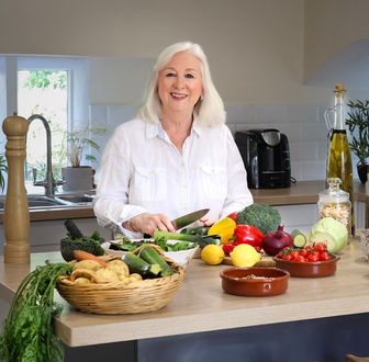 Woman in a kitchen, chopping vegetables; various colorful produce on a counter.