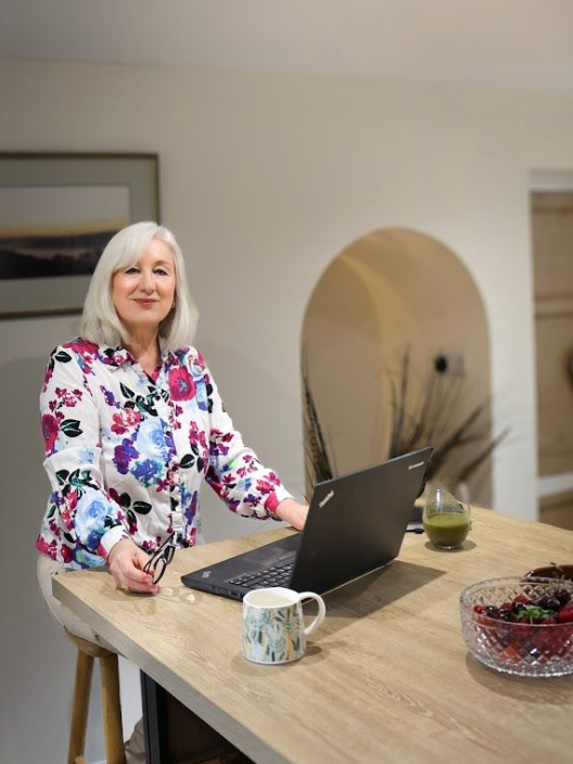 Woman in floral top seated at a kitchen island, working on a laptop, smiling.