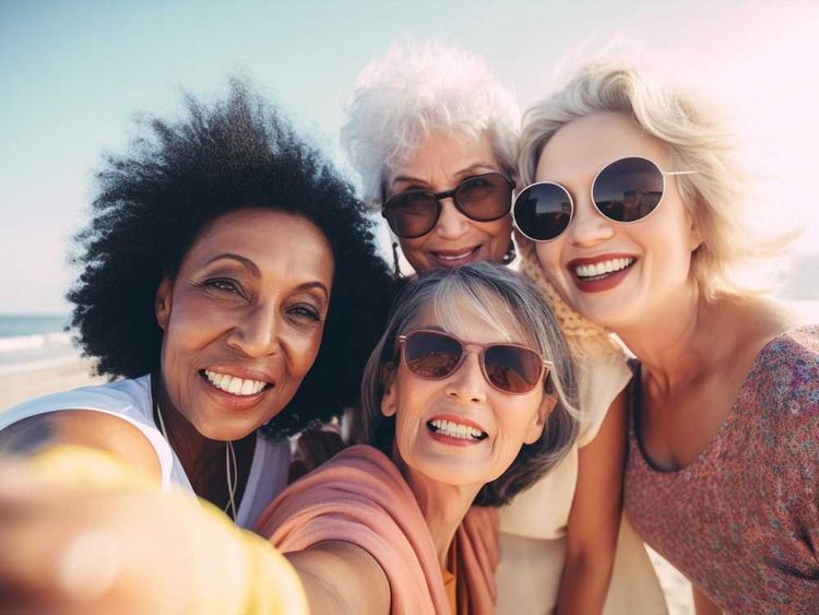 Four diverse women smile, taking a selfie on a sunny beach.