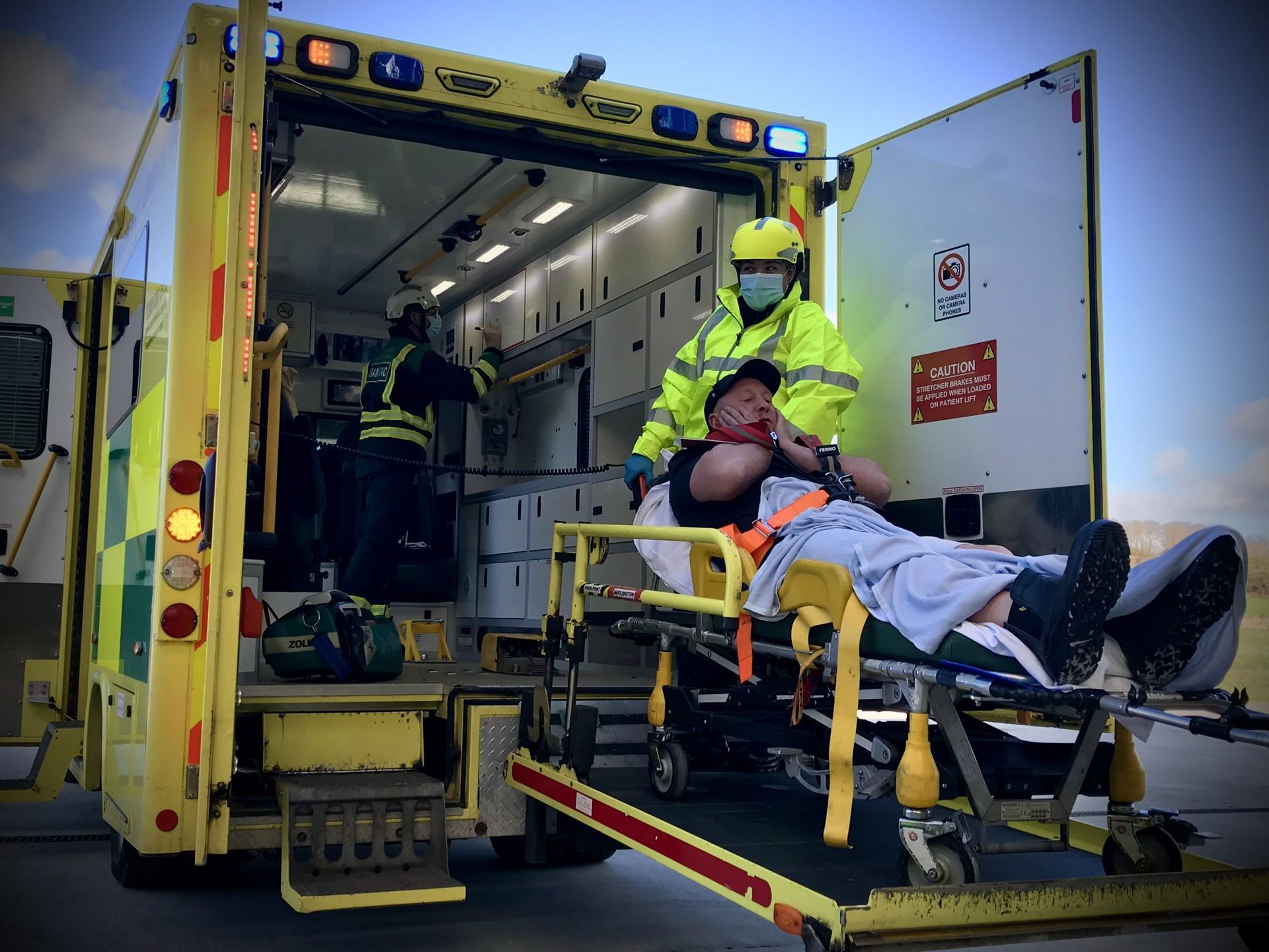 Emergency responders in yellow high-visibility jackets load a patient on a gurney into the back of an ambulance.