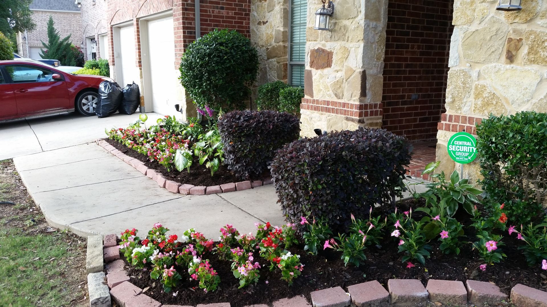 A red car is parked in the driveway of a house with flowers in front of it.