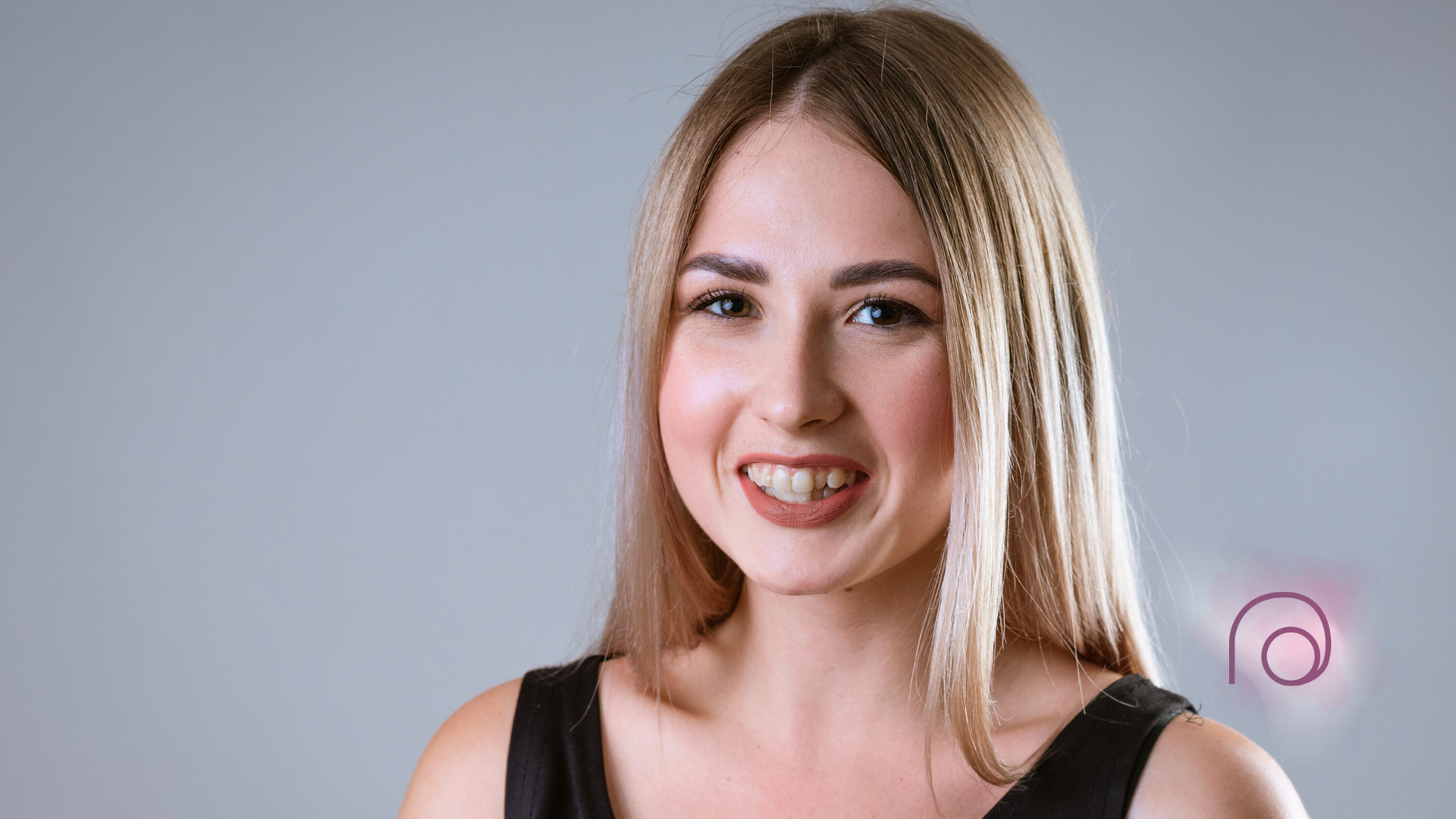 Woman with long blonde hair smiles, wearing a black top. Studio shot, gray background.