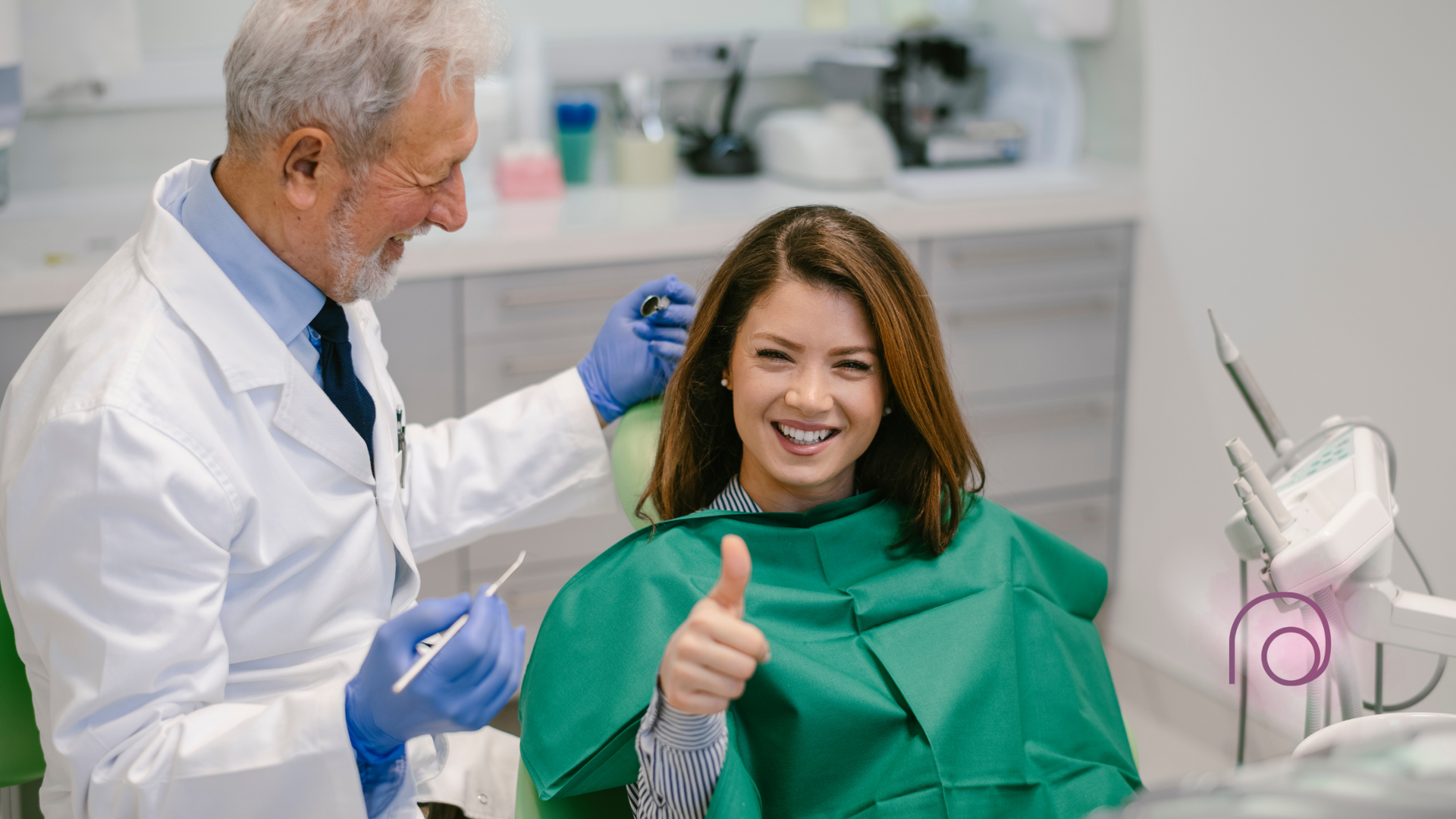 Dentist working on a smiling patient in a green bib giving a thumbs-up in a bright clinic.