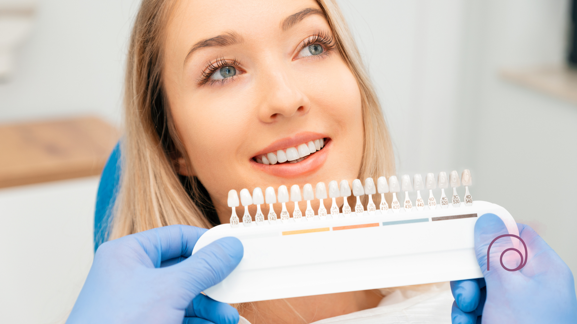A woman is sitting in a dental chair while a dentist holds a tooth color chart.