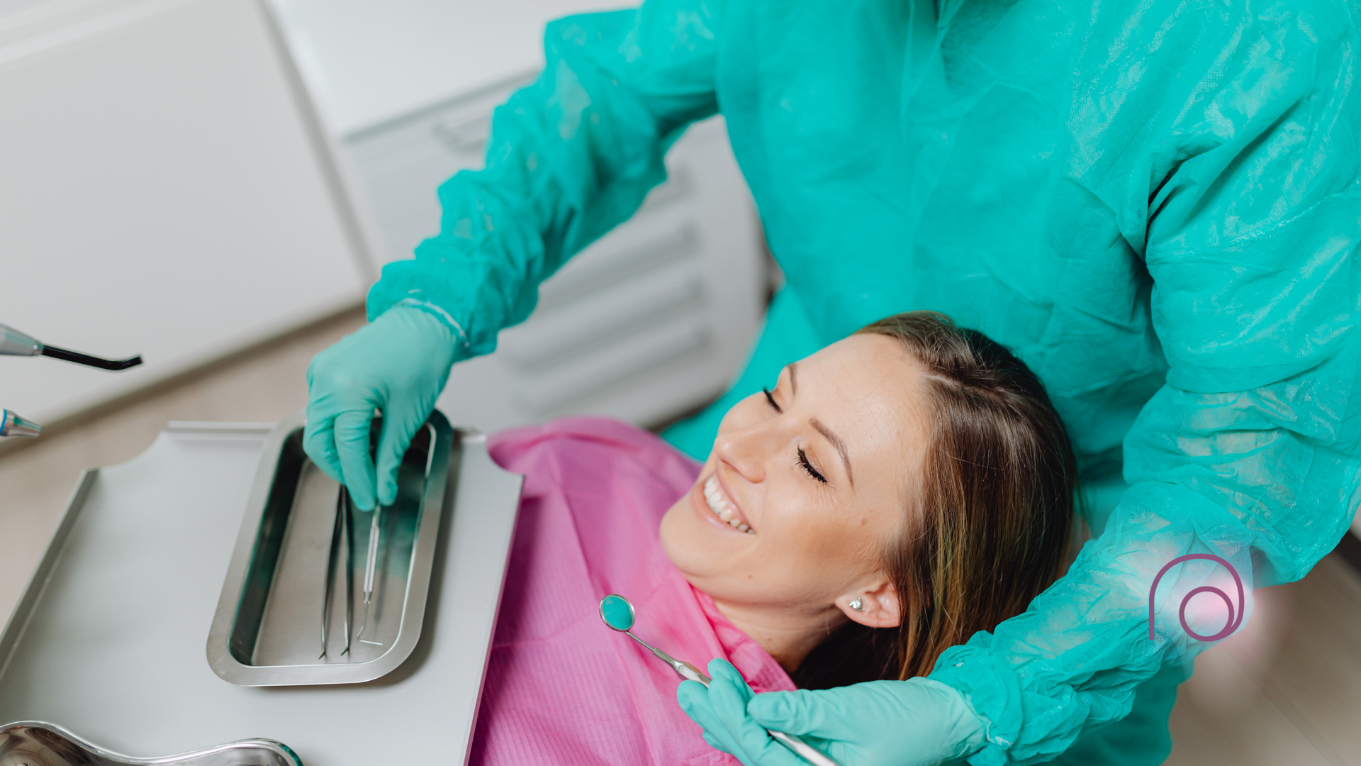Dentist in green scrubs, treating a patient at a dental clinic. The patient is smiling and has a pink bib.
