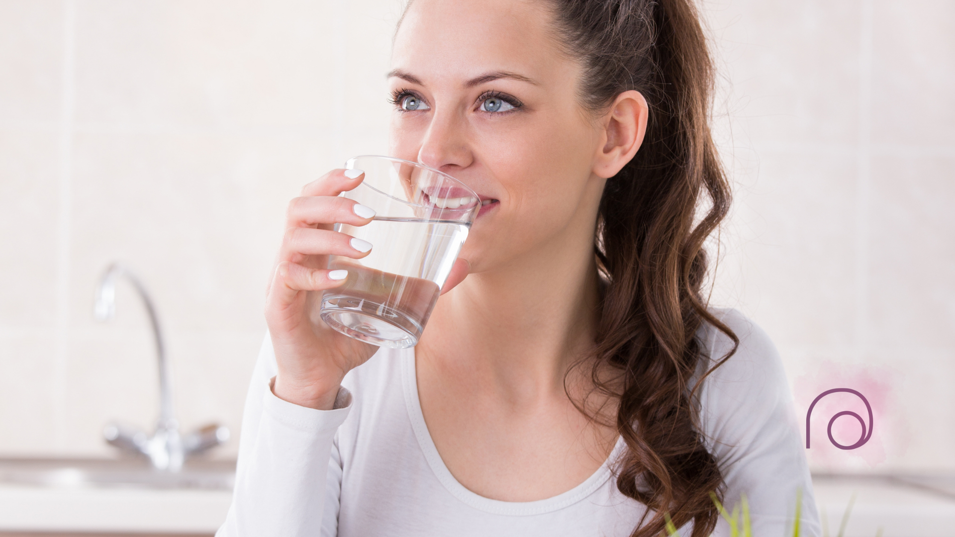 A woman is drinking a glass of water in a kitchen.