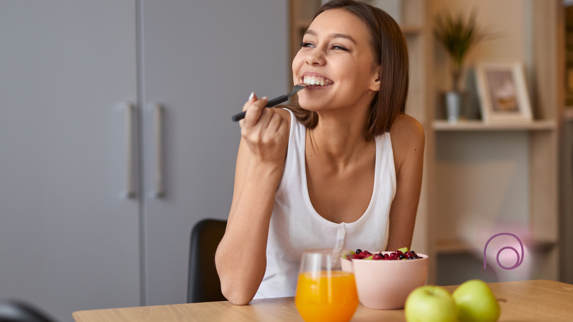Woman smiling while eating breakfast at a table. Orange juice and fruit are visible.