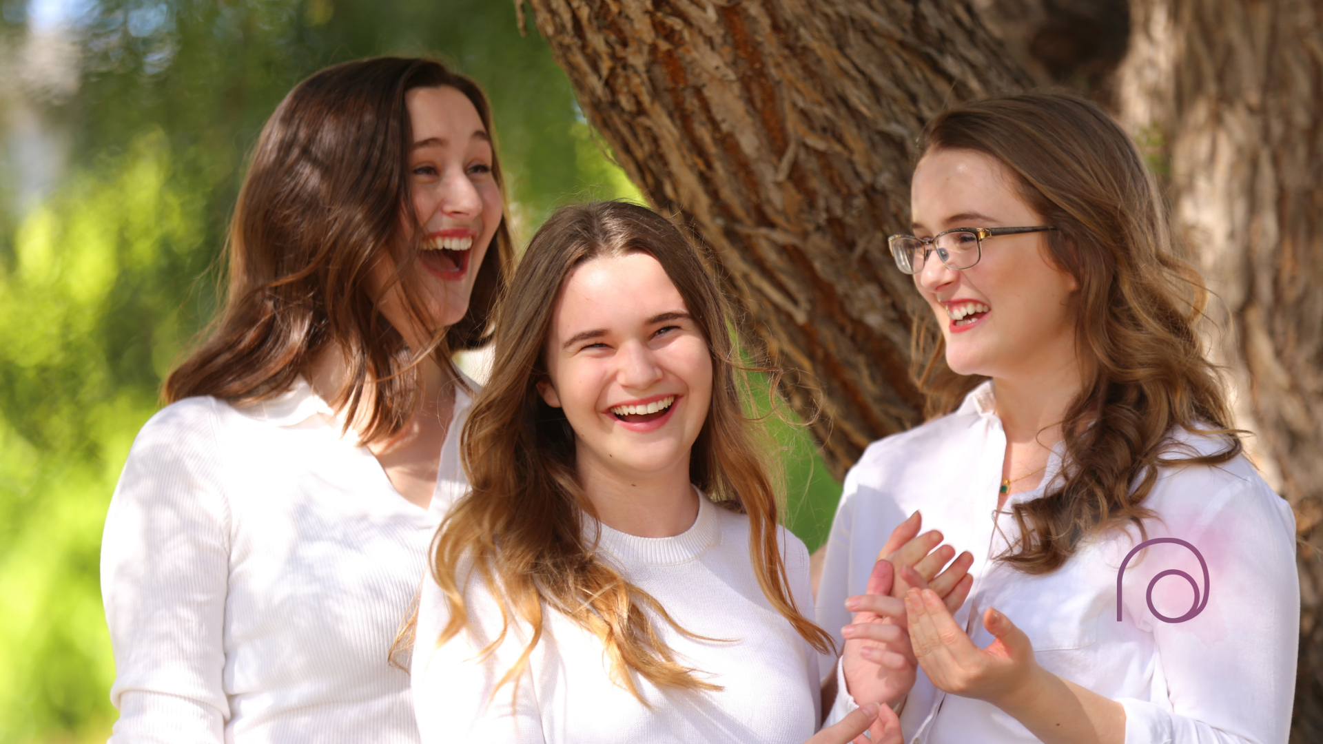 Three smiling people in white shirts outdoors, laughing near a tree.