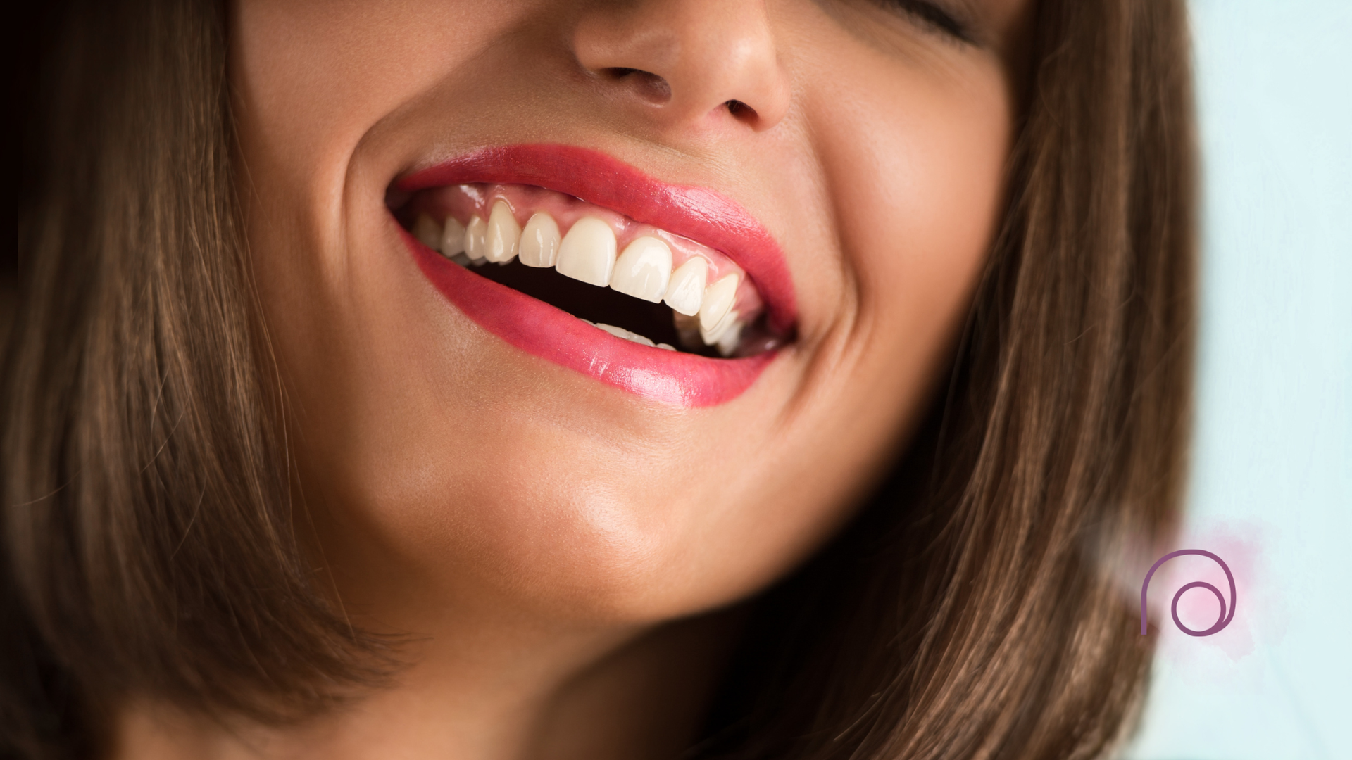 Woman smiling widely, showing white teeth, wearing pink lipstick. Brown hair, light blue background.