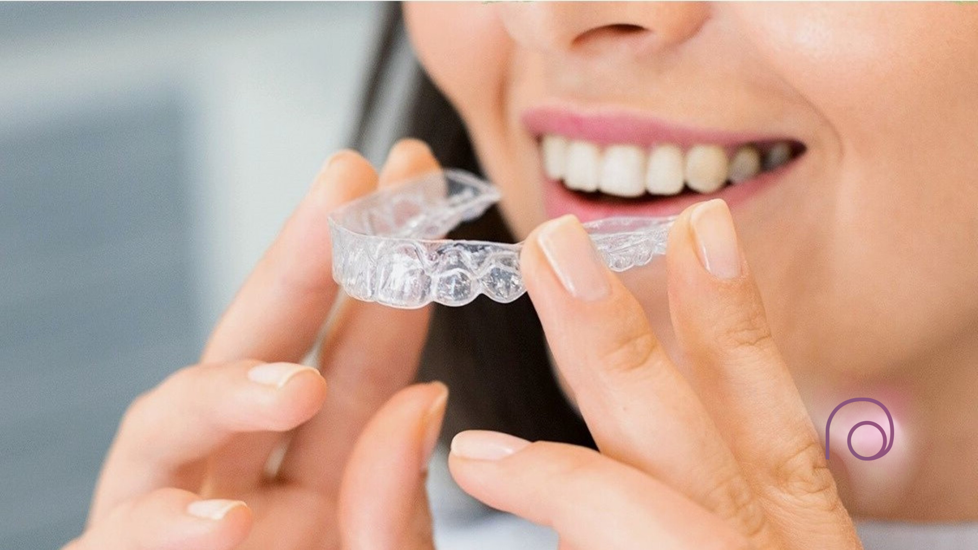 Woman smiling, holding a clear aligner tray near teeth, likely preparing to insert it.