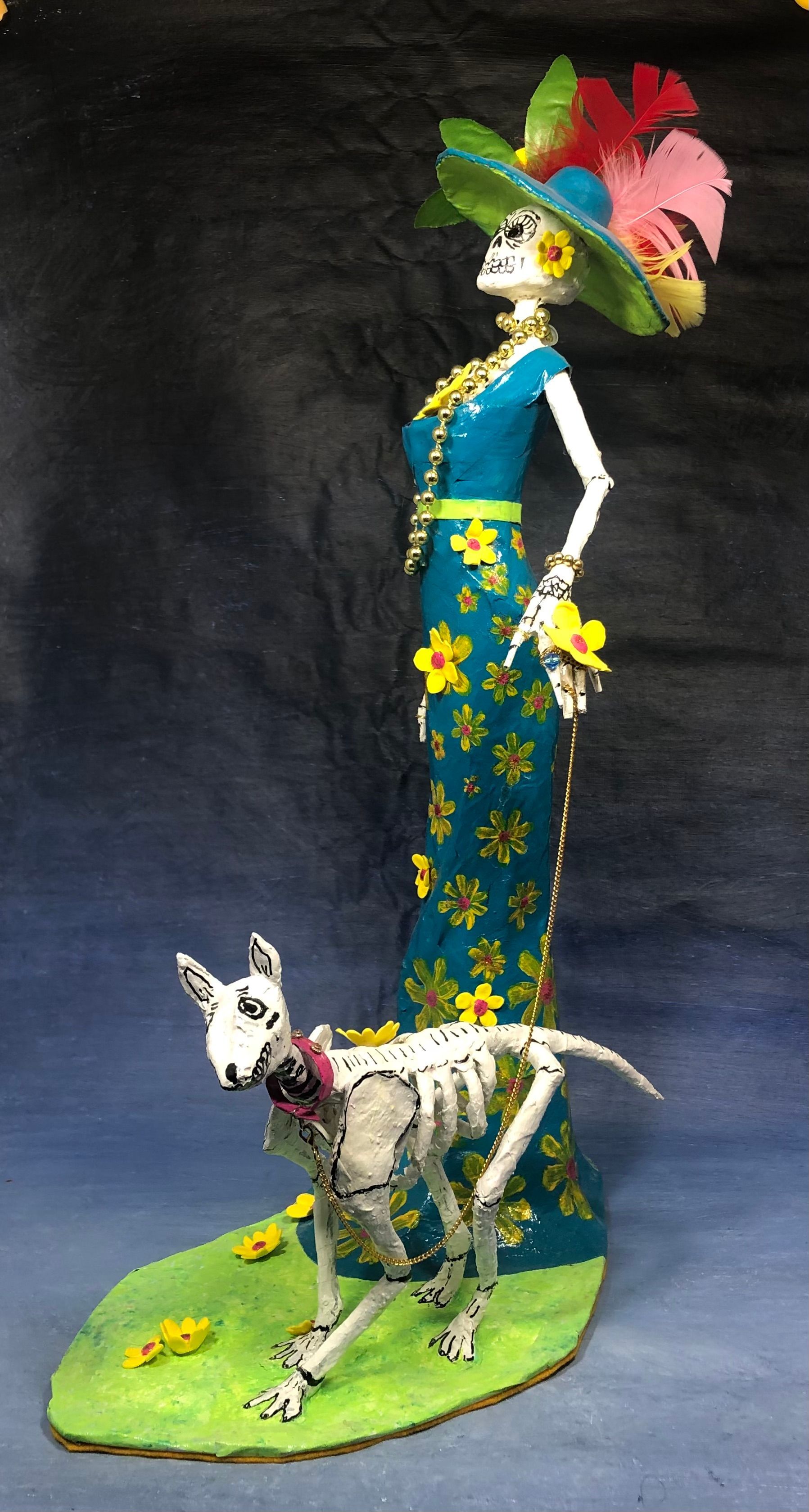 A figuring of a skeleton lady with a wide brimmed feathered hat wearing a blue dress with yellow flowers, walking her dog.