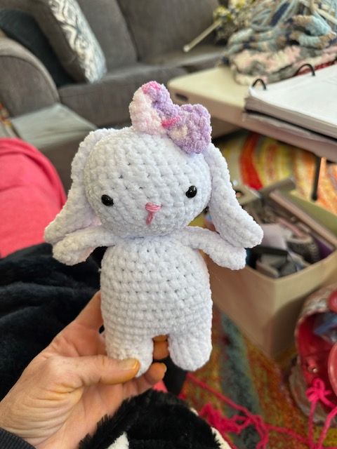 A person is holding a white crocheted bunny with a purple bow on its head.
